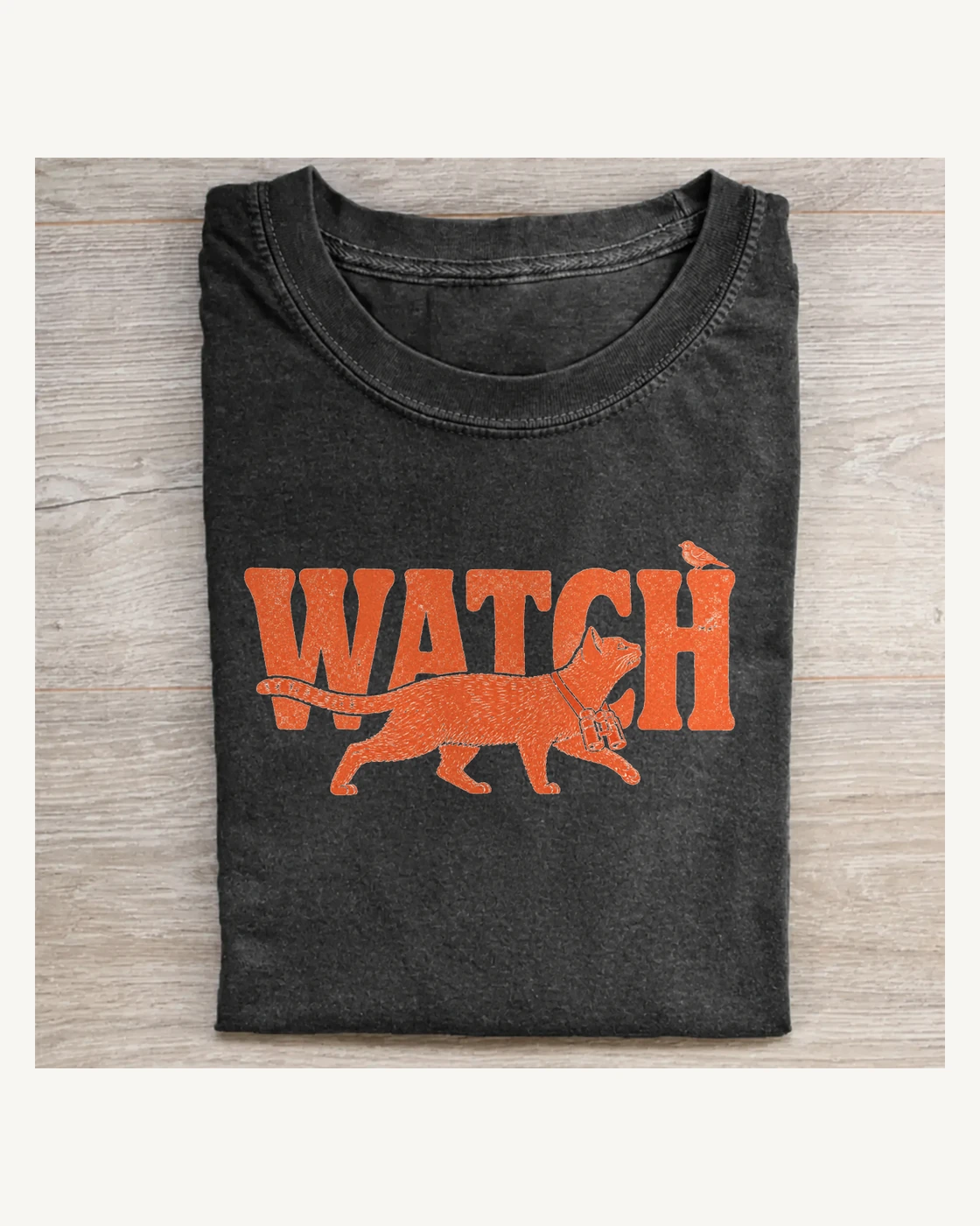 Folded charcoal T-shirt with a burnt-orange binocular cat crossing Watch beneath a tiny bird
